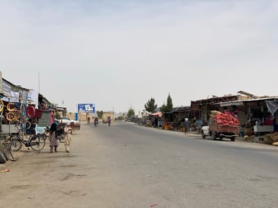 The image shows a street scene in Panjwai, Afghanistan, located at latitude 31.542 and longitude 65.463. The street is paved and lined with small shops on both sides. There is a white pickup truck parked on the right side of the street, loaded with red sacks of what appears to be onions or potatoes.  There are several people walking on the street, including two children, a man in a white turban, and another person in a dark burqa. There are also some motorcycles parked on the side of the road. The sky is overcast with a pale gray hue.  There is a large billboard on the left side of the road that says "15".  The street is relatively empty.