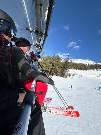 The image depicts a winter ski resort scene at Lunnaya Polyana, Russia, viewed from a transparent-canopied ski chairlift ascending a snowy mountain. In the foreground, three individuals occupy the chairlift. The leftmost person, partially visible, wears a black helmet with reflective goggles and a dark jacket, with a red braided hairstyle visible. Adjacent to them, a male individual with a white beard wears a black knitted beanie, dark sunglasses, a dark ski jacket, and red ski pants. He holds two ski poles in his gloved hands, resting them on the chairlift's safety bar. His red and white "HERO" brand skis are visible below the safety bar, suspended over the snow. A third person, partially visible behind the bearded male, wears a blue helmet and a light blue jacket, also holding ski poles.
The environment below and to the right of the chairlift consists of a wide, groomed ski slope covered in fresh snow, showing ski tracks. Further down the slope, two skiers are visible: one in a red jacket and black pants, and another in a blue jacket and dark pants. The slope is lined with evergreen coniferous trees. In the mid-ground, a red safety net barrier and a yellow marker pole are visible on the edge of the ski run. The background reveals a snow-capped mountain range under a clear, bright blue sky with a few sparse white clouds. The overall scene suggests a sunny day with excellent visibility for skiing.