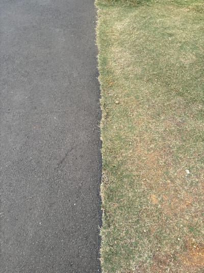 The image shows a view from above of a clear boundary between a dark asphalt surface and a patch of dry grass. The asphalt is textured and appears to be a road or pathway, with a slight sheen suggesting it may be recently laid or slightly damp. The grass is mostly a light brown color with some green patches, indicating it is dry or parched. The transition between the two surfaces is sharp and well-defined, running diagonally across the frame from the top left to the bottom right. No people, animals, or other objects are visible in the scene. The environment appears to be outdoors, with no buildings or man-made structures clearly depicted. The lighting suggests daytime. This image could be a generic depiction of a roadside or park edge in Babakan Madang, Indonesia, or any location with similar landscape features.