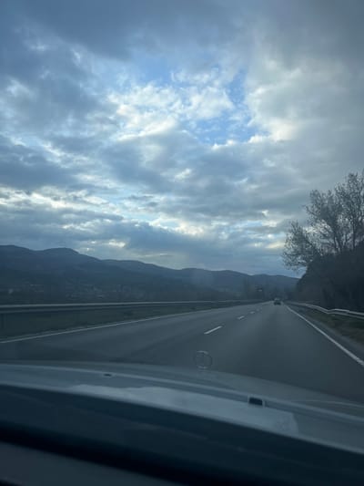 The image depicts a forward-facing view from inside a moving vehicle on a multi-lane highway near Cherniche, Bulgaria. The Mercedes-Benz emblem is visible on the dashboard in the lower center foreground. The highway features at least two lanes of travel in the depicted direction, marked with solid and dashed white lines, and is bordered by metal guardrails on both sides. In the mid-distance, a light-colored sedan travels in the same direction.

To the left of the highway, a series of undulating mountains or large hills extend into the background, showing patches of vegetation and scattered structures consistent with a settlement on their lower slopes. Sunlight penetrates through gaps in the clouds, casting rays onto the distant landscape. To the right of the highway, a steep embankment is covered with trees exhibiting sparse, possibly deciduous, foliage. The sky overhead is heavily overcast with varying shades of gray clouds, but distinct patches of blue sky and bright light are visible, particularly in the upper center, indicating dynamic weather conditions. No individuals are visible. The overall lighting suggests either early morning, late afternoon, or an overcast day with intermittent sun.