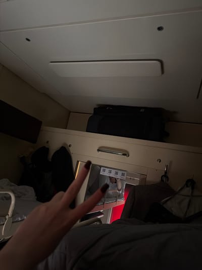 The image depicts the interior of a compact space, likely a train cabin or sleeping compartment, in dim, artificial lighting. The perspective is from inside the compartment, looking towards an internal wall and an adjacent section.

In the immediate foreground, a person's hand, adorned with dark nail polish, makes a "peace" sign (V-gesture) with two fingers extended. Behind the hand, a reflective surface or window reveals a partial view of a person wearing glasses and three slips of paper, each displaying a distinct QR code.

To the left, against a light-colored wall, a dark backpack or bag stands upright on the floor. A narrower, dark rectangular object lies flat on the floor in the foreground, possibly a mat or another item of luggage. On the right side of the frame, a light-colored, built-in panel features a metal handle. From a hook above this panel, a dark-colored, patterned jacket or coat is hanging. A subtle red glow emanates from the background, possibly from an external light source or a window.

The setting, characterized by its utilitarian design and limited space, suggests travel accommodation, consistent with a train journey through a location like Bobrov, Russia. The peace sign gesture conveys a positive or relaxed mood. The visible QR codes are likely related to travel tickets or documents. The scene appears to be taking place at night or in an interior without significant natural light.