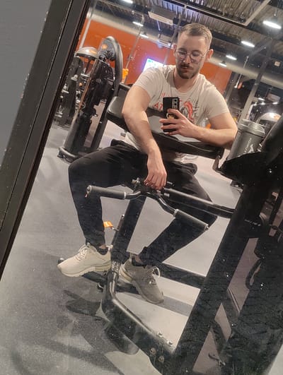 A man is reflected in a mirror at a gym. He is sitting on a piece of exercise equipment, wearing glasses, a white t-shirt, and black sweatpants. He is holding a smartphone in his right hand. A water bottle is sitting on the equipment next to him. The gym equipment and lighting fixtures are visible in the background. There is no specific identifying information to confirm it is the city of Oosterhout, Netherlands.