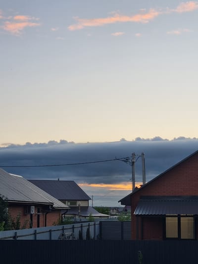 The image depicts a suburban scene in Iskra, Russia, at dusk. The sky is a canvas of soft, pale yellow and grey hues, with a hint of orange from the setting sun visible through a band of dark clouds. Wispy pink clouds are scattered across the lighter part of the sky.
In the foreground and mid-ground, several buildings are visible, their roofs angled towards the viewer. One building on the right is constructed with reddish-brown brick and has a dark grey metal roof. It features a dark-framed window with a soft yellow light emanating from within. To its left, another building with a dark grey metal roof is partially visible. Further into the scene, more houses with grey roofs and various architectural details, like chimneys and air conditioning units, can be seen. A telephone pole with wires stretches across the frame, silhouetted against the sky.
The overall mood is tranquil, characteristic of a quiet evening in a residential area. The time of day is clearly indicated by the colors of the sky and the warm light from the window. The presence of houses and utility poles firmly places the setting in a developed, inhabited area, likely a dacha or residential settlement. No people are visible in the image, and there is no visible text.