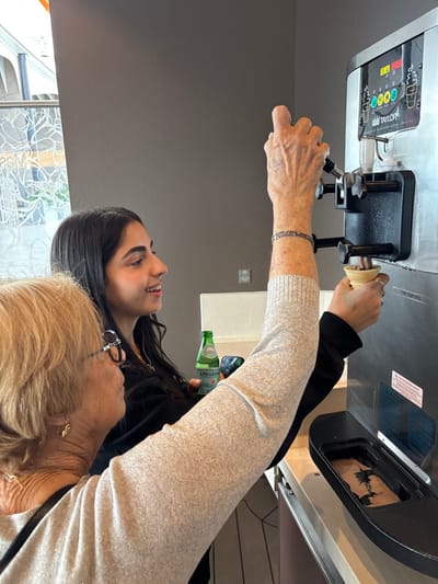 The image depicts two women inside a modern establishment. The precise location within the city of Berry Islands, Bahamas, is not discernible from the image details. An older woman with blonde hair, wearing dark-rimmed glasses and a light-colored long-sleeved top, is operating a stainless steel Taylor soft-serve ice cream machine. Her right arm is extended, manipulating the dispenser lever. She has a multi-strand bracelet on her right wrist and a gold earring and necklace visible. Adjacent to her, a younger woman with long dark hair, wearing a dark top, is positioned in profile, smiling as she holds a brown ice cream cone beneath the dispenser with her left hand. The cone contains a small amount of brown soft-serve ice cream. Her right hand holds a small green glass bottle of S.PELLEGRINO sparkling natural mineral water. The soft-serve machine's control panel is visible, along with a black drip tray below the dispenser, which contains residual brown soft-serve. The background features a plain dark grey wall and large windows on the left with white, abstract decorative patterns. A light-colored countertop runs beneath the soft-serve machine.