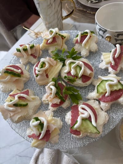 The image shows a close-up, overhead view of a silver-colored, textured glass platter holding approximately a dozen canapés. The canapés are flower-shaped pieces of bread topped with what appears to be salami or ham, cucumber slices, cheese slices, and a white sauce, possibly mayonnaise or a similar dressing. Fresh parsley sprigs are interspersed between the canapés. Part of a white teacup with black cat silhouettes is visible in the upper-left corner. The platter rests on a white surface, potentially a table, with hints of a golden utensil and other tabletop items visible in the periphery. There is no indication of Nottingham, United Kingdom.