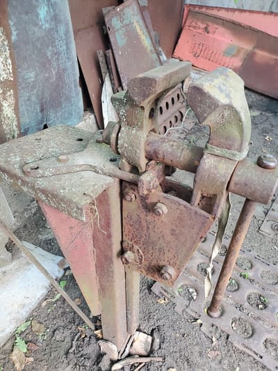 The image depicts a heavily rusted vise mounted on a metal workbench. The workbench itself appears to be made of thick, weathered metal plates. The vise is in a state of disrepair, with visible signs of corrosion and wear. It is surrounded by other pieces of scrap metal and debris, suggesting an outdoor storage or work area. The ground is a mix of soil and gravel, with some scattered leaves. The overall scene is one of neglect and disuse. The location is identified as Konstantinovka, Moldova.