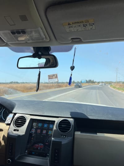 An interior view from a vehicle shows a dashboard with an aftermarket infotainment system, a rearview mirror with an air freshener hanging, and a sun visor with airbag warning labels. The exterior view through the windshield reveals a road with a yellow line, sparse vegetation on either side, and a few vehicles in the distance. A billboard is visible on the right side of the road, beneath a clear blue sky.