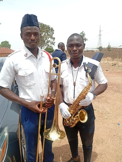 This outdoor photograph features two African men in uniform, prominently displaying musical instruments under a bright, hazy sky. They appear to be members of a band or a uniformed organization.
The man on the left wears a white, short-sleeved collared shirt adorned with a red cord over his right shoulder and a crest insignia on his collar, which seems to include a cross and an anchor. He sports a dark blue garrison cap and dark blue trousers. He holds a large, golden brass trombone vertically, with its bell resting on the ground. A grey car is partially visible to his left.
The man on the right wears a similar white, short-sleeved collared shirt with a crest insignia, black trousers, and white gloves. A dark cap or ceremonial shoulder piece is secured over his right shoulder, and he wears a distinctive bolo tie. He holds a gleaming golden saxophone, its bell pointed upwards. Both men look towards the camera, with the man on the right displaying a slight smile.
The setting is an open, dry, and dusty unpaved area. In the background, there are scattered low-rise buildings, some featuring corrugated iron roofs, alongside sparse green trees. A prominent metal power pylon stands in the distance on the right, indicating an electrical grid. The bright, hazy sky suggests it is daytime. The overall environment, with its distinctive architecture and infrastructure, points to the provided location context of Ladura Foron, Nigeria. The men’s attire and instruments suggest they are either about to perform or have just concluded an event.