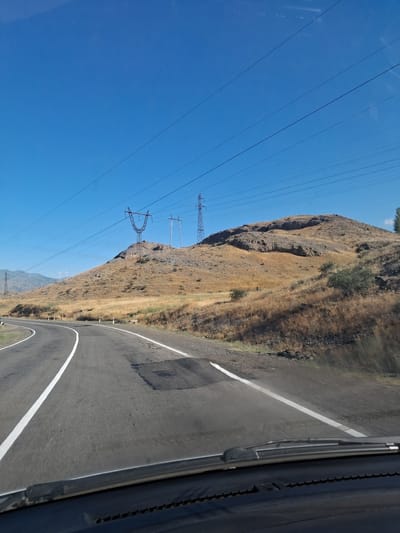 A road curves through a dry, grassy hillside under a clear blue sky. Power lines stretch across the upper portion of the frame, supported by tall, lattice-like towers, several of which are visible on the hillside and in the distance. The road has white lane markings, and a small patch of darker asphalt is visible in the right lane. The scene is viewed from inside a vehicle, with the dashboard and windshield wipers visible in the foreground. The location is identified as the vicinity of Malishka, Armenia.