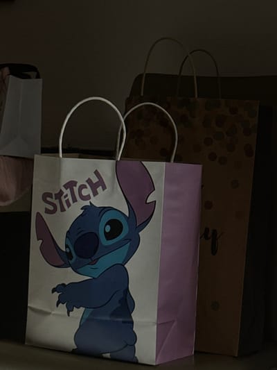 This scene, situated in the city of Albi, France, depicts an indoor, dimly lit environment featuring multiple paper gift bags arranged on a flat surface. In the immediate foreground, a white and light purple gift bag stands upright. This bag features a prominent graphic of the blue Disney character Stitch, with large ears, and the word "Stitch" printed in purple above its head. To the right of and slightly behind this primary bag, a taller, darker brown gift bag is visible. It is patterned with numerous dark circular dots and exhibits partial black cursive text, specifically the letter "y," on its lower right side. To the far left, a portion of another white gift bag and an indistinct reddish-pink or light brown object are partially visible in shadow. No persons are present within the frame, and no actions or interactions are depicted beyond the static arrangement of the objects.