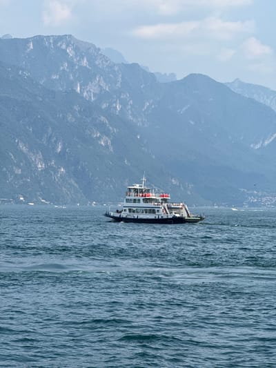 This media file is a photograph of a ferry on a large body of water with mountains in the background. The ferry is white with multiple decks and railings, and it appears to be moving. There are some people visible on the deck of the ferry, but they are small and not clearly distinguishable. The water is choppy, indicating some wind. The mountains are large and imposing, covered in vegetation, and shrouded in a slight haze, suggesting either a humid or polluted atmosphere, or simply distance. The sky is partially cloudy. The scene is bathed in bright light, implying it is daytime, likely late morning or afternoon. The overall mood is serene, capturing a moment of travel and natural beauty. The photograph's orientation is vertical, with the water occupying the right side and the mountains the left. No visible text is present in the image. The location context of Loveno, Italy, places this scene on one of Italy's famous lakes, likely Lake Como, given the characteristic landscape of mountains meeting water.
