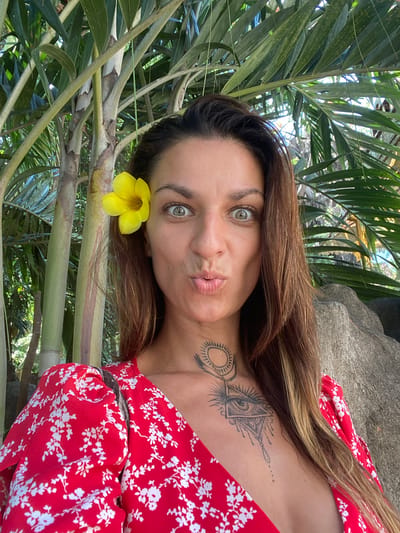 A woman is pictured outdoors, among tropical foliage. A yellow flower is placed in her hair near her ear. She is wearing a red dress with a white floral pattern. Tattoos are visible on her neck and chest. The location is unspecified, and there is no indication it is in Dĩ An, Vietnam.