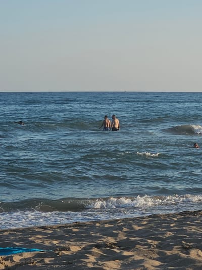 This is a photograph taken from the beach in Castelldefels, Spain. The scene depicts the Mediterranean Sea with gentle waves breaking on the shore. In the middle ground of the water, two adult males are standing, appearing to be in conversation. To their right, a third person, possibly a child, is visible in the water, with only their head above the surface. Further to the left, another person is swimming in the sea, and a small sailboat is visible on the distant horizon. The foreground of the image shows a sandy beach with a blue towel laid out. The sky is clear and pale, indicating fair weather.