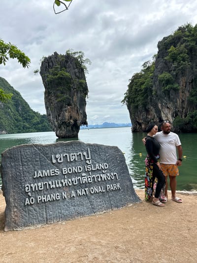 A couple stands in front of a stone monument at James Bond Island, in Krasom, Thailand. The man, wearing a white t-shirt and orange patterned shorts, has his arm around the woman. She is wearing a black long-sleeved shirt, black pants, and a colorful patterned wrap. They are both looking to their left. Behind them, a distinctive limestone karst, known as James Bond Island, rises out of the turquoise water. The sky is overcast, and lush green vegetation covers the surrounding cliffs. The stone monument in the foreground has text in both Thai and English, identifying the location as "JAMES BOND ISLAND" and "AO PHANG NGA NATIONAL PARK". The ground is sandy.