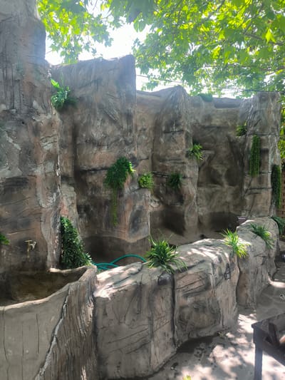 The image shows a manufactured rock structure with simulated crevices and ledges. Artificial plants are interspersed throughout the structure. A faucet is visible on one section. A green hose rests on the top surface of the lower section. The structure appears to be outdoors, as indicated by the sunlight and foliage overhead. The image location is identified as Karatakla, Georgia.