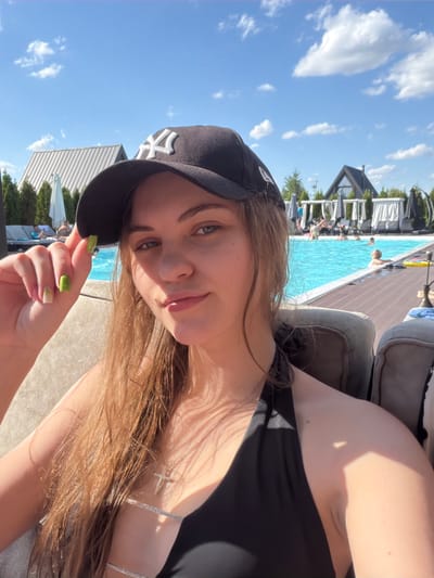 In the city of Slobozhanske, Ukraine, a young woman with light brown hair and a black baseball cap is captured in a selfie. She is wearing a black bikini top and a silver cross necklace. Her hand, adorned with bright green nail polish, rests on the brim of her cap. Behind her, a swimming pool shimmers under a clear blue sky dotted with white clouds. The poolside is furnished with lounge chairs and umbrellas, and several people are visible in and around the water. The background reveals modern architectural structures, suggesting a resort or a well-equipped residential complex. The sunlight casts shadows, indicating it is a bright, sunny day.