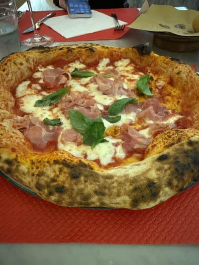 A close-up, top-down view of a freshly baked pizza, likely Neapolitan style, is presented. The pizza has a puffy, charred crust and is topped with tomato sauce, melted mozzarella cheese, thin slices of ham, and fresh green basil leaves. It rests on a dark green serving platter, which is placed on a textured red surface.

To the left of the pizza, a white paper napkin is visible, folded in a triangular shape. A silver fork is partially resting on the napkin. Further to the left, a small smartphone with a blue case is visible, displaying a grid of colorful app icons. A clear glass wine glass is also present, positioned in front of the napkin and fork, with its stem and base catching the light. In the background, a small portion of a brown, round object is seen, possibly a serving dish or a part of the kitchen equipment.

The scene appears to be indoors, given the lighting and the arrangement of objects, suggesting a casual dining setting, perhaps a restaurant or a home kitchen. The pizza's appearance, with its slightly irregular shape and artisanal crust, points to it being handmade. The presence of fresh ingredients like basil indicates a focus on quality and presentation. The overall impression is one of a delicious, ready-to-be-enjoyed meal. There is no visible text in the image. The context provided places this scene in Bergamo, Italy.