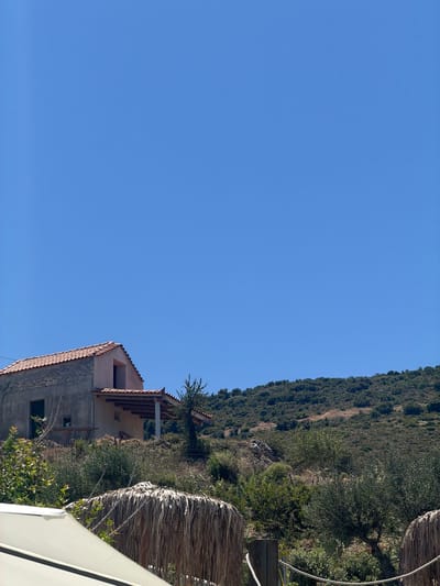 The image depicts a building with a tiled roof, possibly a house or a guesthouse, situated on a hillside in Elaties Municipal Unit, Greece. The building appears to be under construction or renovation, with unfinished sections visible. Dense vegetation, including trees and shrubs, covers the steep slope behind the structure. The scene is bathed in bright sunlight, suggesting a clear day.

In the foreground, two thatched umbrellas or sunshades are partially visible, indicating an outdoor seating or relaxation area, possibly associated with a hospitality venue. A white awning or canopy is also present in the lower right corner, further suggesting a tourist or recreational setting.

The image captures a tranquil, sun-drenched atmosphere. The hillside location and the presence of Mediterranean vegetation, such as olive trees, hint at a rural or semi-rural setting within Greece. The clear blue sky and the quality of light suggest it is daytime, likely late morning or early afternoon. There is no visible text in the image.