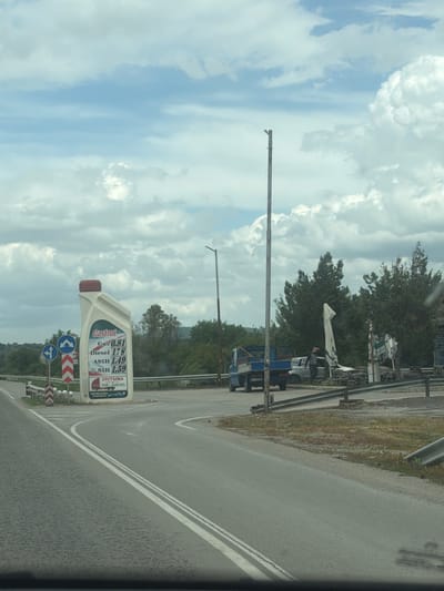 The image shows a road in Belomortsi, Bulgaria, with a large sign shaped like a Castrol oil bottle displaying fuel prices: Gas 0.81, Diesel 1.78, A95H 1.49, and 95H 1.59. In the background, there's a blue truck parked near trees and other roadside structures. The sky is cloudy. Road signs indicating directions are also visible.