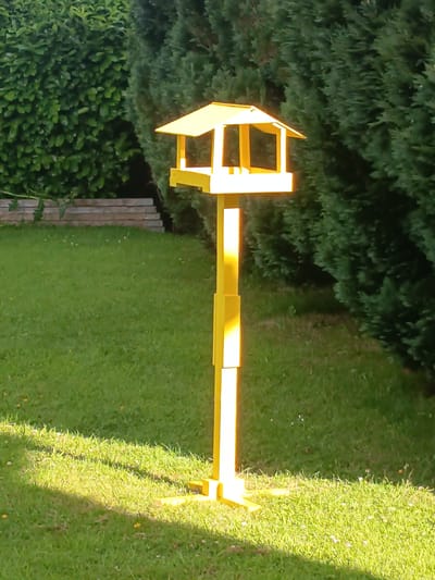 A bright yellow bird feeder stands on a lawn in Millstreet, Ireland. The feeder is constructed of wood and has a pitched roof with open sides. It is supported by a tall, thin yellow post which widens at the base to form a stable footing. The lawn is well-maintained, with green grass and scattered white wildflowers. In the background, a dense hedge of dark green foliage forms a backdrop. The scene is bathed in natural sunlight, casting shadows on the grass.