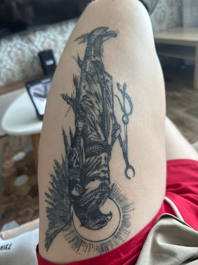 A black and grey tattoo of an Egyptian deity, possibly Anubis or Thoth, is depicted on a person's thigh. The figure is shown upside down, with a jackal head and human body adorned with intricate clothing and jewelry. The tattoo includes a crescent moon with rays emanating from it, and a winged headdress. The person is wearing red shorts. The background is out of focus but suggests an indoor setting with furniture and a table. The image was taken in the city of Kondopoga, Russia.