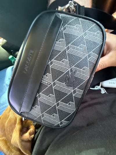The image shows a close-up view of a black and white geometric pattern toiletry bag with the "LACOSTE" logo embossed on a black leather strip. The bag has a small, dark embossed crocodile logo on its patterned surface. A person's hand is visible on the right side of the image, holding the bag. The bag is placed on a dark fabric surface, possibly clothing, and a small part of a brown dog is visible at the bottom left. In the background, a partially visible green bottle is present. There is no indication of the city of Vyzyrka, Ukraine, in the image.
