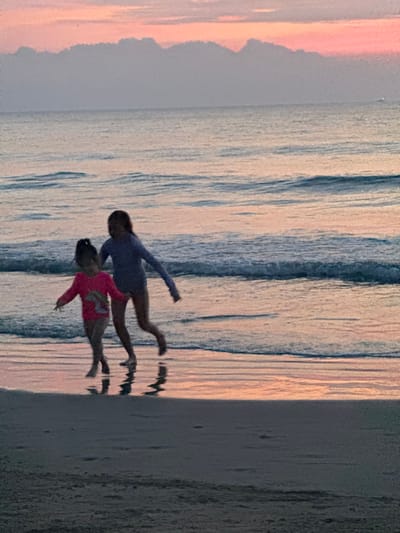 Two figures, one in pink and the other in blue, appear to be suspended in air or water. They are positioned above a body of water, possibly the ocean, with a pale sky above reflecting on the water's surface. To the right, a vertical sandy stretch meets the water. The scene is illuminated with a soft light, creating a serene and dreamlike atmosphere.