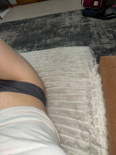 The image shows a close-up view of a person's leg and torso, wearing grey underwear and a white t-shirt. The skin on the upper thigh exhibits stretch marks. The person is lying on a textured, light-colored blanket or comforter, which is placed on top of a rug with a dark grey and black pattern. To the right of the blanket, a brown, textured object, possibly a piece of furniture or a cushion, is visible. In the background, on the rug, there are two backpacks, one maroon with a white logo and the other dark blue. The environment appears to be indoors, possibly a room in a home. There is no discernible information to locate the scene in Postoyata, Ukraine.