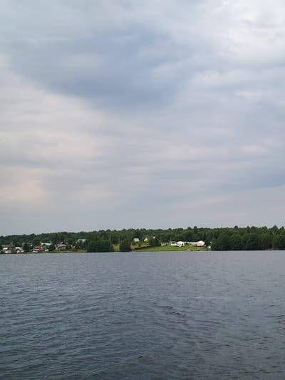 The image depicts a wide, dark blue body of water in the foreground, with small ripples and waves on its surface. In the middle ground, a tree-lined shore is visible, with a small village nestled among the trees. The village consists of several houses with red and brown roofs, and some buildings with white roofs. Further along the shore, there are several white domed structures, possibly tents or small buildings, situated on a grassy area. The background is filled with a dense forest of green trees, which extends across the horizon. The sky above is overcast, with a mix of light gray and darker gray clouds, suggesting a cloudy day. The scene appears to be a view from a boat or the edge of the water, looking towards the opposite shore of a lake or river in Kameshnitsa, Russia.
