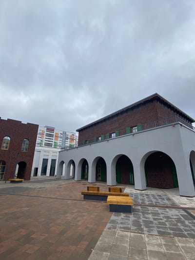 This is an outdoor shot of an urban scene in Chemoshur, Russia. The buildings are characterized by white arches and brown brickwork, with some having green accents. The pavement is made of brown and grey tiles. There are wooden benches. The sky is overcast with grey clouds. No people are visible.