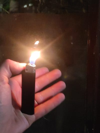 A hand holds a black lighter with a flame burning at the top. The lighter is dark, with some metallic accents around the flame. The flame is bright white and yellow, with a hint of blue at the base. The hand is light-skinned and appears to be holding the lighter firmly. The background is dark and blurry, with some hints of green and brown, suggesting an outdoor setting at night, possibly in a garden or wooded area. The overall lighting is dim, with the lighter providing the primary light source. The scene is likely captured in Makhinjauri, Georgia, given the context of the request.