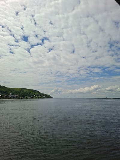 The image depicts a wide, calm river under a partly cloudy sky. In the distance, a verdant, gently sloping hill is visible, dotted with small buildings and trees. The buildings appear to be houses, some with red roofs, clustered along the shoreline. The river extends towards the horizon, where the faint outlines of more distant buildings can be seen, suggesting a town or city on the opposite bank. The water is dark and reflects the sky and the surrounding landscape. The clouds are scattered and varied in shape and density, with patches of blue sky visible between them. This scene is likely a view of the Volga River near Verkhny Uslon, Russia, a settlement known for its picturesque riverside location.