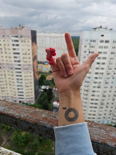 A first-person perspective shot is taken from above, focusing on a left hand with two fingers extended in a "peace" sign. A red plastic toy adorns the index finger, resembling a miniature clown or puppet. The hand is positioned in front of a backdrop of high-rise buildings, suggesting an urban environment. The sky is overcast with gray clouds. A rusted metal railing runs along the bottom of the frame, and a portion of a light blue garment is visible on the lower left. On the wrist, a tattoo of a circular aperture is visible, with some Japanese characters above it. The location is identified as Novoivanovskoe, Russia.