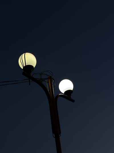 The image shows a lamppost with two spherical lights against a dark blue sky. The lamppost is made of dark metal and has a curved design. The top light is bright and white, while the bottom light is slightly dimmer and has some horizontal lines on it. Wires are visible connecting the lights to the lamppost. The image's location, Ogradna, Bulgaria, is not indicated in the photograph.
