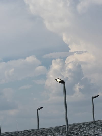 This media file shows a view looking upwards towards a cloudy sky with the presence of streetlights and a large metal structure.

**Subjects:** The primary subjects are three streetlights mounted on metal poles extending from a large, grid-like metal structure. The streetlights are modern, rectangular in design, with one appearing to be illuminated. The metal structure resembles scaffolding or a large fence, possibly part of a stadium or industrial facility. The background is filled with a cloudy sky, suggesting an outdoor setting.

**Activity or Event:** No specific activity or event is depicted. The image captures a static scene, likely a moment in time outdoors.

**Notable Details:**
*   **Time of Day:** The lighting conditions suggest it is daytime, possibly late afternoon or early evening, given the diffuse light and the illuminated streetlight, which might be testing or pre-dawn/post-dusk.
*   **Weather:** The sky is heavily clouded, indicating overcast or partly cloudy weather.
*   **Location Cues:** The metadata indicates the location is Singen (Hohentwiel), Germany. This context, combined with the industrial or stadium-like structure, could hint at a public space or venue.
*   **Emotions:** The image evokes a sense of a neutral or perhaps slightly somber mood due to the overcast sky and the utilitarian appearance of the structures.

**Visible Text:** There is no discernible text in the image.