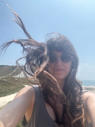 A selfie is taken by a woman with long, dark, wavy hair blowing in the wind. She is wearing sunglasses and a dark grey tank top. She is standing outdoors with a bright blue sky behind her. In the background, there is a scenic view of rolling green hills with a path winding through them, and the sea in the distance. The image appears to be taken on the South Downs Way near Seaford, East Sussex, United Kingdom.