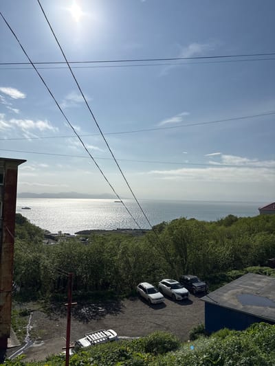 In a medium shot, a bright sun shines through a clear blue sky with scattered clouds, casting shimmering reflections on a vast body of water. Power lines crisscross the frame, originating from a weathered, reddish-brown building on the left. In the foreground, a dirt parking area is visible, with three SUVs parked: a white SUV on the far left, a white SUV in the middle, and a dark SUV on the right. Behind the parking area, lush green trees form a dense forest that extends to the water's edge. The water is dotted with several boats, and distant mountains form a hazy horizon. On the far right, a blue building with a dark, flat roof and a large puddle on its surface is partially visible. A white vehicle with a roof rack is parked near the bottom of the frame, to the left of the blue building, partially obscured by greenery. The overall impression is a coastal town scene in Vulkanyy, Russia, on a sunny day.