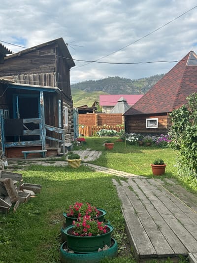 The image depicts a rural yard in the village of Yabogan, Russia, under a cloudy sky. A weathered wooden house with a blue-painted porch and door is prominently featured on the left. In the foreground, a pathway constructed from wooden planks winds through the grassy yard. Several potted plants, including vibrant pink petunias in green containers, are visible. Further into the yard, a wooden fence encloses a garden area with various flowers and some vegetables. A second, smaller wooden building with a red-tiled roof is situated behind the fence. In the background, rolling green hills with scattered trees rise against the sky. The overall impression is one of a simple, lived-in rural setting.