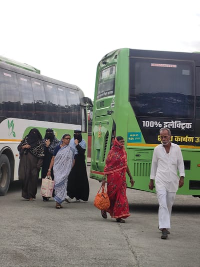 The image shows a group of people standing and walking on an asphalt surface in front of two buses. The bus on the left is white and green with a leaf logo. The bus on the right is green and has "EMERGENCY EXIT" marked in red on the window. The bus on the right also has text in Hindi. A man in a white outfit walks away from the camera towards the right. A woman in a red and floral sari walks past him to the left, carrying an orange bag. A group of four women, three of whom are wearing hijabs, stand near the left bus. One of the women is carrying a light-colored bag. The scene appears to be a bus station or a similar transportation hub in Datala, India, given the Hindi text on the bus and the presence of traditional Indian attire. The overall atmosphere is neutral and functional.
