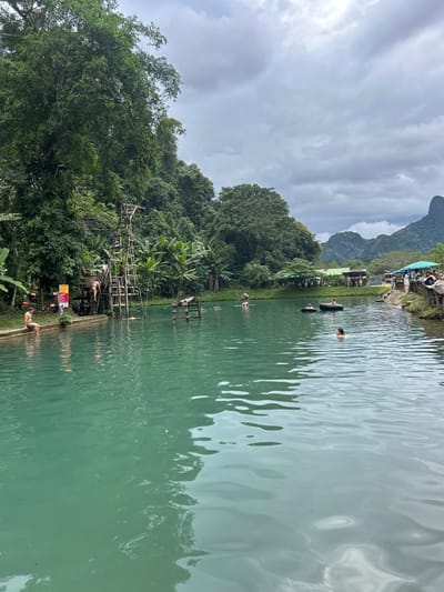 The scene is an outdoor recreational area located in Ban Nampoun, Laos, featuring a large, still body of turquoise water under an overcast, cloudy sky. The left bank is characterized by dense, tropical foliage including large trees and banana plants, and supports a multi-tiered wooden diving platform. One individual in dark swimwear is ascending the platform, while another in light swim trunks sits on a concrete edge at the water's immediate left. A smaller, partially submerged wooden structure is present in the water near the left bank. In the mid-ground water, one individual is swimming, and two others occupy separate black inflatable tubes. Additional individuals are partially visible in the water further back. The right bank features several simple structures, including those with blue tarpaulin roofs, and a gathering of approximately 10-15 people on a wooden platform. Distant karst mountains frame the background.