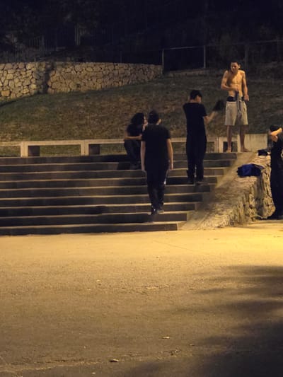 A group of young men are gathered on a set of concrete stairs at night. The man at the top of the stairs is shirtless and wearing shorts. Another man is standing next to him, holding a shirt. Two other men are on the stairs, wearing black shirts and pants. The environment appears to be an outdoor urban setting with a stone wall and grassy embankment behind the stairs. The scene is lit by artificial lights. It is implied that this scene is taking place in Cornellà de Llobregat, Spain.