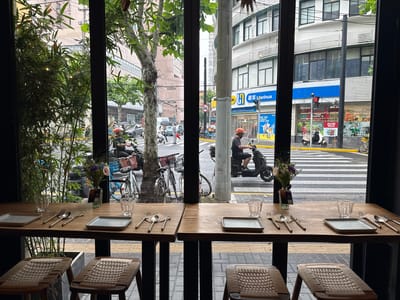 The image presents an interior view from a restaurant or cafe, looking out onto a lively urban street in Jing'an District, China. In the foreground, two wooden counter tables are set with square, light grey plates, gold-colored cutlery (forks, spoons, knives), and clear glasses. Small vases holding flowers and small signs labeled "B1" and "B2" are also on the tables. Woven-seat stools are pulled up to the counters, suggesting a casual dining or drinking spot. A large potted bamboo plant frames the left side of the window view.
Through the tall, paneled windows, a street scene unfolds under what appears to be overcast daylight. Numerous individuals on scooters or mopeds, cyclists, and pedestrians traverse the road and zebra crossing. One person on a scooter, wearing an orange helmet, is centrally visible, while another person with an umbrella crosses the street, hinting at recent rain or drizzle. Bicycles are parked along the pavement.
In the background, multi-story urban buildings line the street. A prominent storefront displays the characters "联华" alongside the English "Lianhua," and the slogan "美好生活由此进 HAVE A GOOD DAY." Another partially visible sign reads "IVE THE GOOD LIFE." Advertisements for "可口可乐" (Coca-Cola) with the text "此刻" (This Moment) and "一口透心凉" (One sip, refreshing to the heart) are visible on a shop window. A red traffic light is active, and a 'No Entry' or similar regulatory road sign is also present. The scene depicts a typical bustling city street, viewed from a tranquil indoor setting.