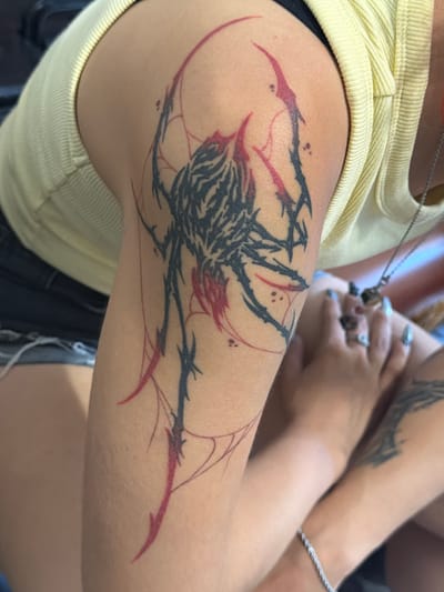 The image shows a close-up of a person's arm with a black and red spider tattoo. The tattoo is abstract and stylized, with sharp lines and a tribal aesthetic. The person is wearing a yellow ribbed tank top and denim shorts. Their hands are visible in the foreground, with manicured nails and rings on their fingers. The background is blurred, but suggests an indoor setting. There are no discernible landmarks or features that would indicate the city of Hostalric, Spain.