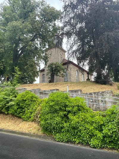 The image shows a stone church with a clock tower situated on a grassy hill, surrounded by lush green trees. The church features arched windows and a red-tiled roof. A stone cross stands on the hill near the church. In the foreground, there are dense green bushes and a paved road. The scene appears to be in a rural or suburban setting, likely in the city of Hattendorf, Germany, given the architectural style and vegetation. The sky is overcast, suggesting a cloudy day. There are no people visible in the image.