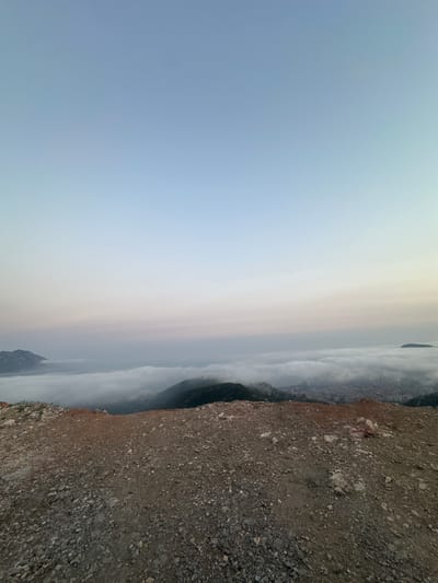 An aerial, wide-angle shot captures a misty dawn over a coastal city nestled amongst mountains. The foreground features a rough, rocky terrain with reddish-brown dirt and scattered stones, suggesting a construction site or undeveloped land. In the middle ground, a thick blanket of white fog obscures a city and its surrounding hills, with only the tops of some mountains and buildings emerging from the mist. The distant background reveals a calm sea meeting a pale blue sky, transitioning into lighter hues near the horizon, indicating the early morning light. There are no people or animals visible in the image. The location is identified as Markovići, Montenegro.