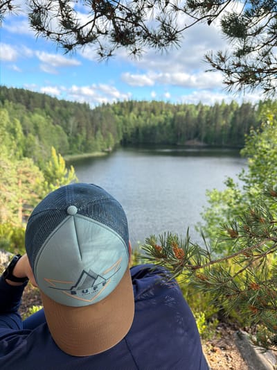 The image shows a person wearing a dark blue shirt and a light blue and brown cap, looking out at a body of water. The person is positioned in the foreground, with their back to the camera, leaning forward slightly. The cap has a mesh back and a light blue front panel with a logo that appears to be "BUFF SINCE 1992" along with a mountain graphic. The background features a serene lake surrounded by a dense forest of green trees. The sky above is blue with scattered white clouds. Pine branches with small, developing cones are in the foreground, framing the view of the lake and forest. The lighting suggests it is daytime, with sunlight filtering through the trees. The overall scene depicts a moment of quiet contemplation in a natural setting. The location is identified as Ruch'i, Russia.