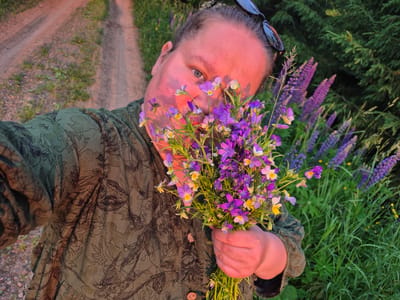 A person, viewed from a high angle, is holding a bouquet of wildflowers in their right hand. They are wearing a green shirt with a dark leaf pattern. Sunglasses are perched on their head. The person's face is partially obscured by the flowers, and their skin is illuminated by warm, pinkish light. A dirt road runs through the background, flanked by green grass and tall purple lupine flowers. The scene appears to be in a rural or natural setting, possibly in or around the city of Dojnaya, Russia.
