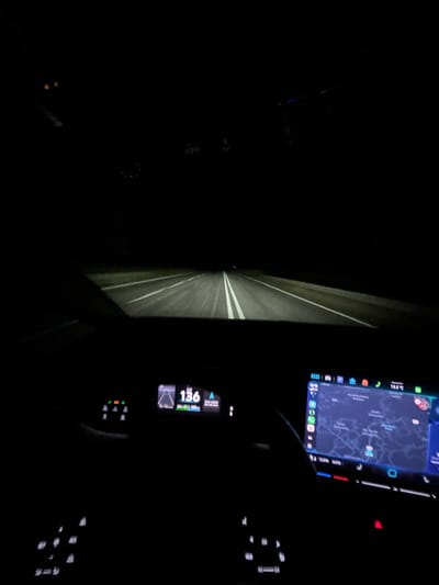 The image shows the interior of a car at night, with the headlights illuminating a dark, empty road. The driver's perspective is forward, looking out the windshield.
On the dashboard, there are two screens. The screen on the left displays a digital speedometer showing "136" km/h, along with other vehicle information. The screen on the right is a navigation system displaying a map, indicating the car is in or near the city of Amarante, Portugal. The map shows roads and building outlines.
The road ahead has white lane markings that are illuminated by the car's headlights. The environment outside the car is dark, with no other vehicles or significant landmarks visible. The interior of the car is dimly lit, with the screens and some dashboard lights being the primary sources of illumination.