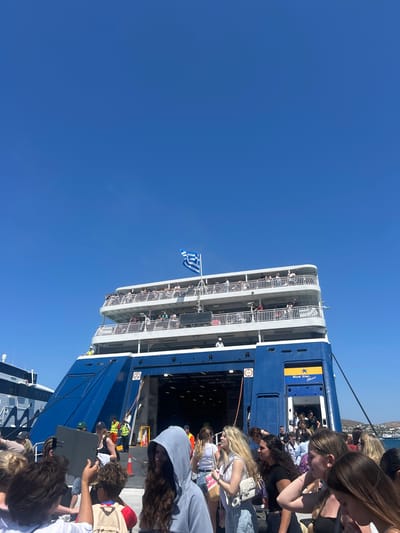 In Parikia, Greece, a blue and white ferry, "Blue Star Naxos," is docked at the port under a clear blue sky. The ferry has multiple decks with white railings, and passengers are visible on these decks, some standing and looking out. A Greek flag flies from the top deck. Below, a ramp leads into the ferry's open cargo bay. A crowd of people, appearing to be tourists, are gathered near the ramp and on the dock. Some are carrying bags and appear to be embarking or disembarking. In the background, other boats and buildings are visible on the waterfront. Two individuals in yellow vests are positioned near the ramp, seemingly directing passengers. The overall scene depicts a busy port environment on a sunny day.