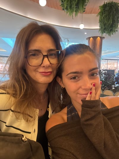 Two women are posing closely together for a selfie indoors. The woman in the foreground has shoulder-length brown hair, eyeglasses, and is wearing a light-colored, striped sweater. Behind her, the other woman has her hair pulled back, is wearing a brown sweater, and has red nail polish. The background includes indoor foliage, decorative lighting, and reflections of storefronts. This location is likely a shopping center in Senhora da Hora, Portugal.